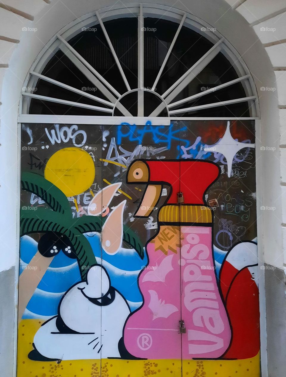 Street art of graffiti. Image of a person(apparently a vampire) sunbathing on a sand beach, with a sunscreen bottle on the front; a palm tree, the sun and the sea also can be seen