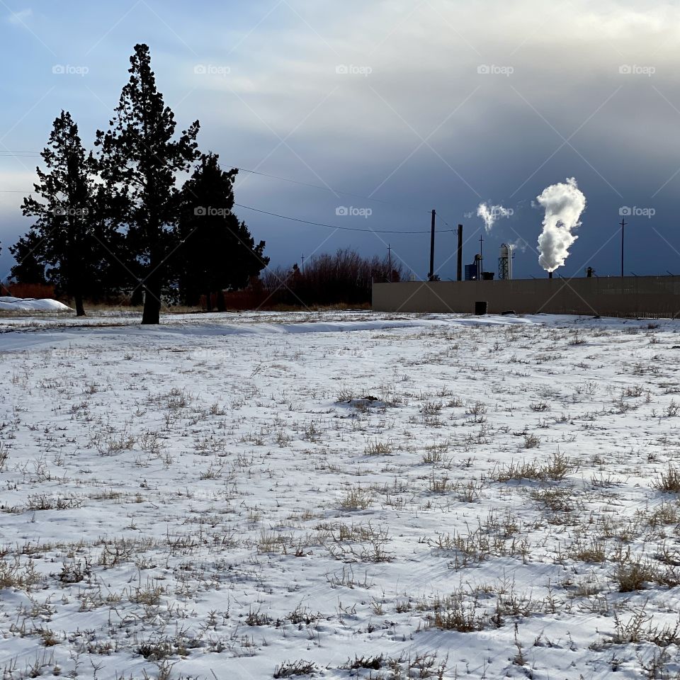 Bright white steam from an industrial plant at the edge of a snow covered field contrasts against a dark and stormy sky. 