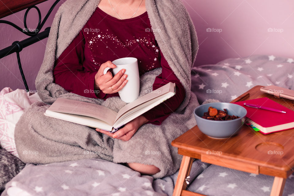 Woman enjoying the reading a book and drinking coffee at home. Young woman sitting in bed, wrapped in blanket, holding book, relaxing at home. Candid people, real moments, authentic situations