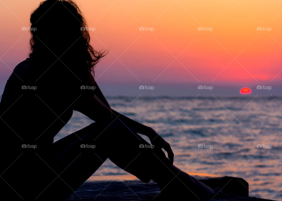 peaceful relaxed silhouette of woman watching a pinkish orange sunset over sparkling blue water