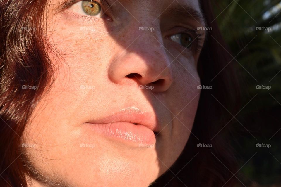 Women with natural and minimal makeup standing outdoors outside with wet hair. Women with red hair and green eyes standing next to tree taking a selfie. Sunlight shine on women hair and skin and making shadows. Women wearing a hat.