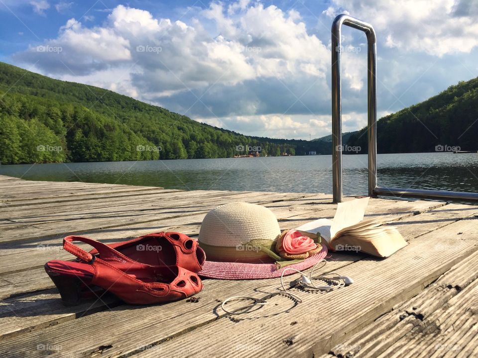 summer items on wooden deck: red sandals, hat, book and headphones near the lake