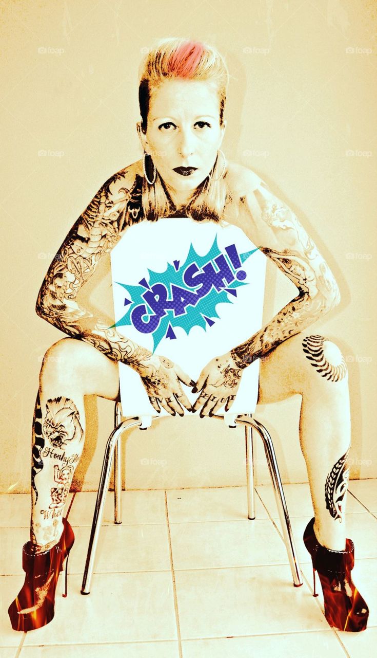 Tattooed Blonde Model in Chair , showing both tattooed arm sleeves and the legs , but covering her entire torso with a white chair , with the word crash in dark blue and teal blue exaggerated onto the back of the white chair . The words evil blood across her fingers & skull and cross bones on left hand , orange koi fish on her right hand . Hot closed black boot heels are appearing to be on fire . The picture is in two tones , the chair and word crash are in color and the model and back ground of the picture are in a sepia tone  .