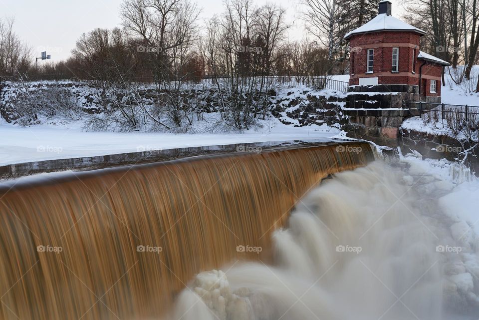 Helsinki, Finland - March 10, 2021: Water pours over frozen museum dam in the mouth of Vantaa River at the Vanhankaupunginkoski rapids (Vanhankaupunginkosken putous) on extremely cold winter morning.