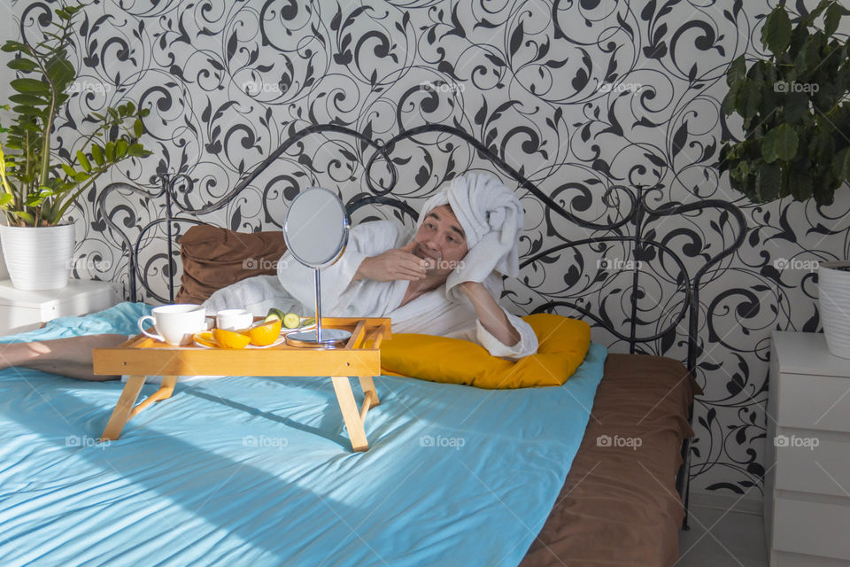 A man and home spa care at home takes care of himself and smears his face with cream, does a manicure at home on a blue background and with oranges and rests.